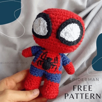 Una mano sostiene un muñeco de crochet de Spider-Man con ojos grandes y exagerados, un traje rojo y azul y un emblema de araña negro en el pecho. El texto de la imagen dice "PATRÓN GRATIS DE SPIDERMAN", con un pequeño identificador de Instagram "@CROCHET.BY.MAYA" visible en el fondo.