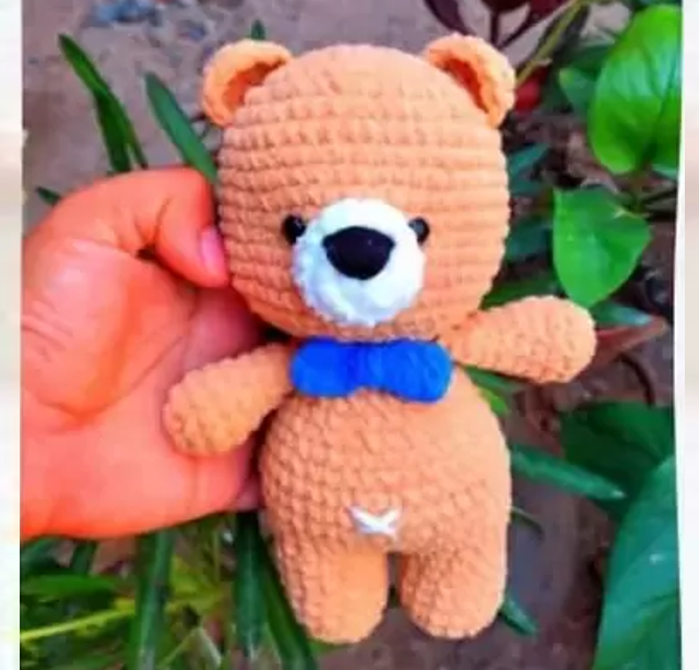 Un Osito De Peluche Está Sostenido Por Una Mano El Oso Que Recuerda a Un Oso De Peluche Amigurumi Es De Color Marrón Claro Con Un Hocico Blanco Ojos Negros Nariz Negra Y Una Pajarita Azul Alrededor Del Cuello El Fondo Muestra Plantas Verdes Y Un Escenario Al Aire Libre Borroso