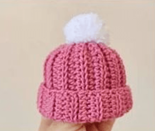 Una Mano Sostiene Un Gorro De Bebé De Punto Rosa Con Un Pompón Blanco Y Esponjoso Encima Sobre Un Fondo Liso Perfecto Para Quienes Dominan El Crochet Para Principiantes