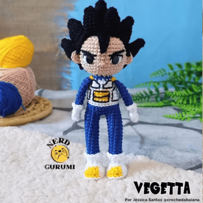 Una muñeca de ganchillo que se asemeja a un personaje animado se encuentra sobre una superficie texturizada con un fondo borroso. El amigurumi Vegeta tiene cabello negro puntiagudo, un traje azul y blanco con un emblema amarillo y zapatos con puntas amarillas. El texto dice "NERD GURUMI" y "VEGETTA" de Jéssica Santos @crochedabaiana.